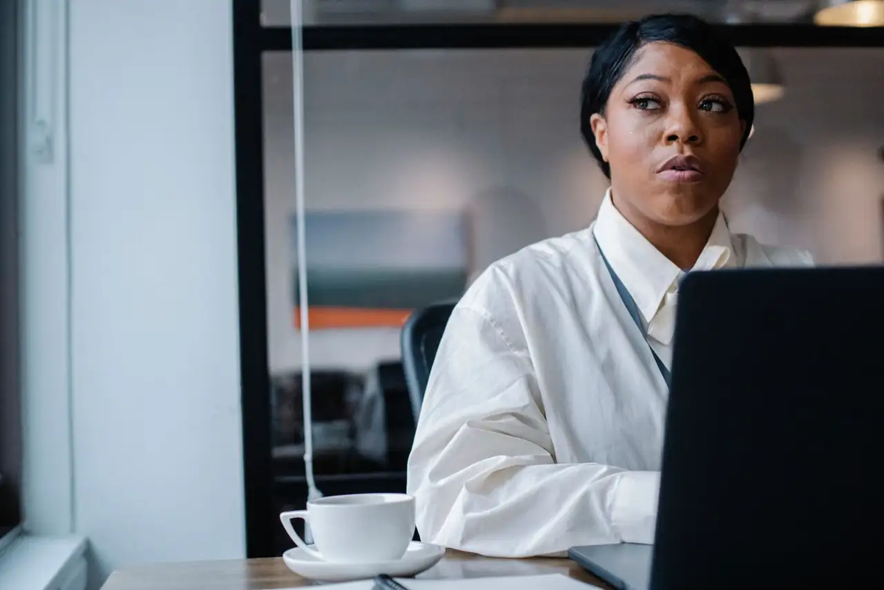 Serious African American female employee wearing white formal shirt sitting at working desk with laptop and cup of coffee